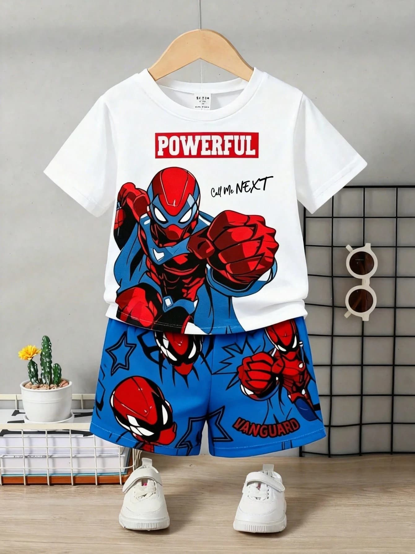 A kids' two-piece outfit on a hanger — a white 'POWERFUL' superhero T-shirt with matching blue comic-print shorts, styled with white trainers and round sunglasses.