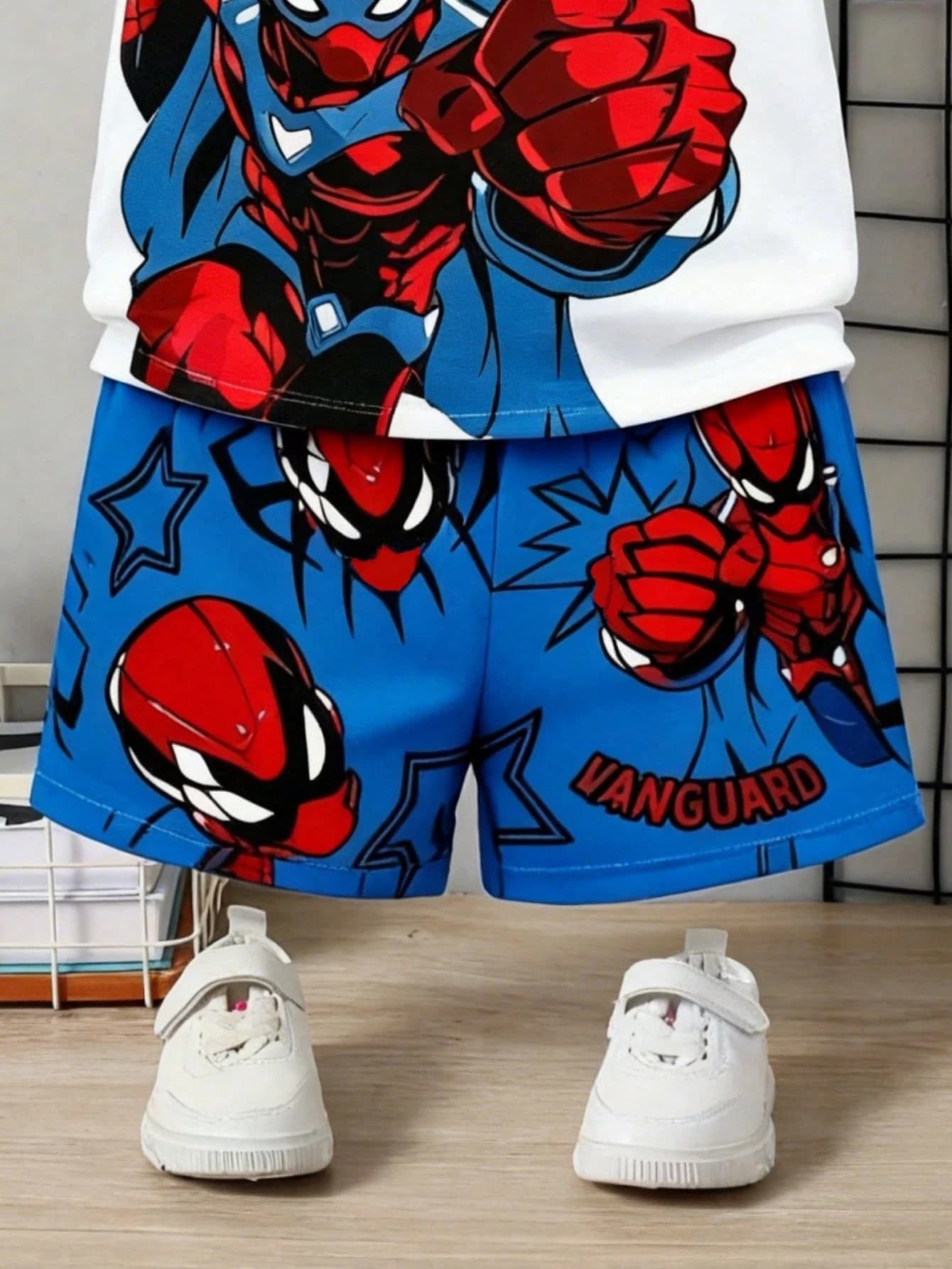 Close-up of the kids' outfit shorts — bright blue with a red-and-black comic superhero print reading 'VANGUARD' — above white trainers.