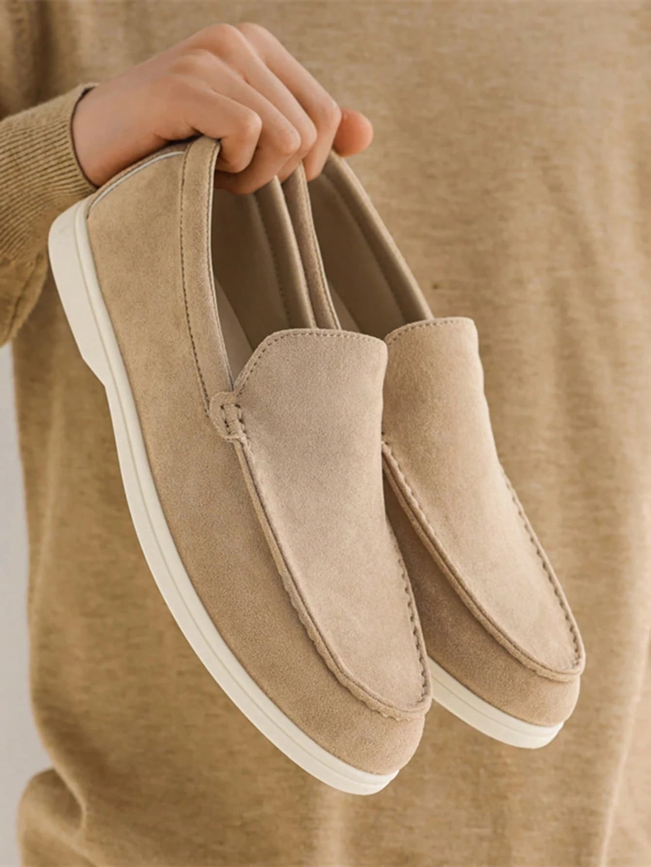 A pair of beige suede slip-on loafers with cream soles, photographed on a wooden floor.