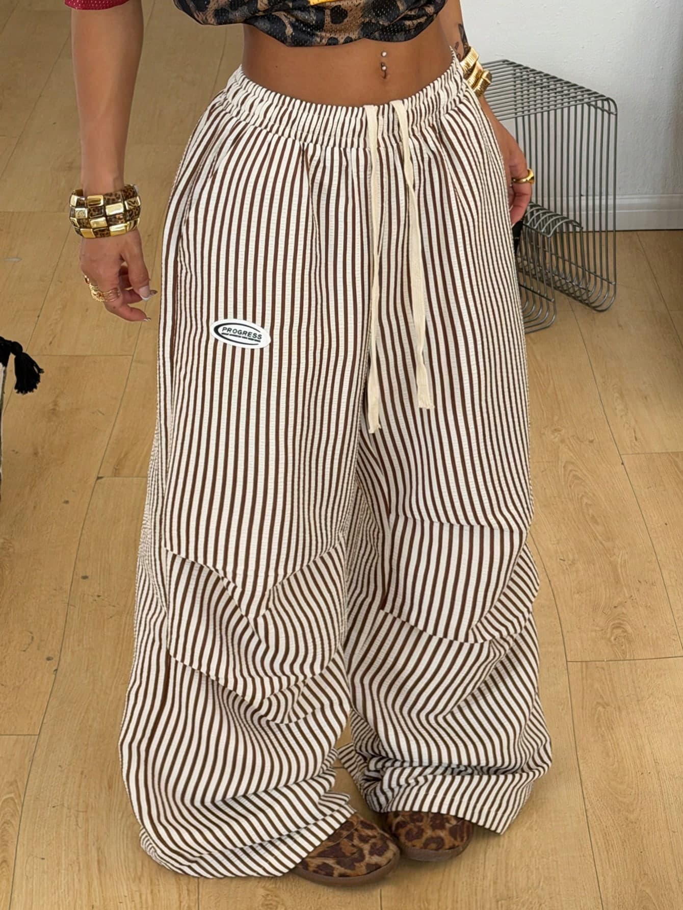Brown-and-cream vertically striped wide-leg cargo pants worn with a cream drawstring waist and a small oval PROGRESS patch on the thigh, photographed on a wooden floor.